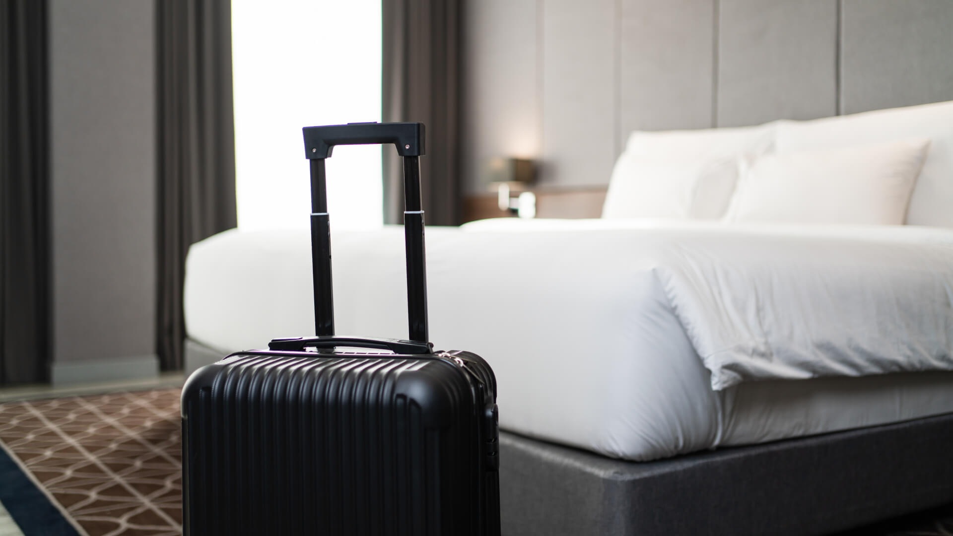 Black suitcase in front of a neatly made hotel bed.