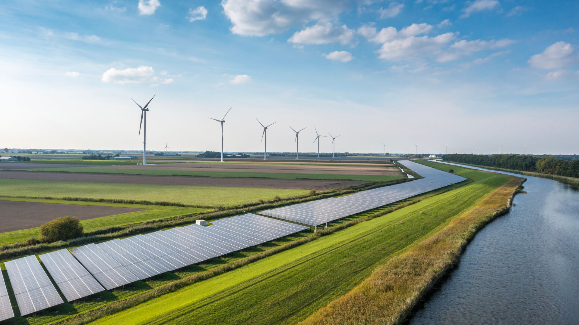 Aerial view of solar panels and wind turbines in a green rural landscape beside a wide canal under a blue sky.
