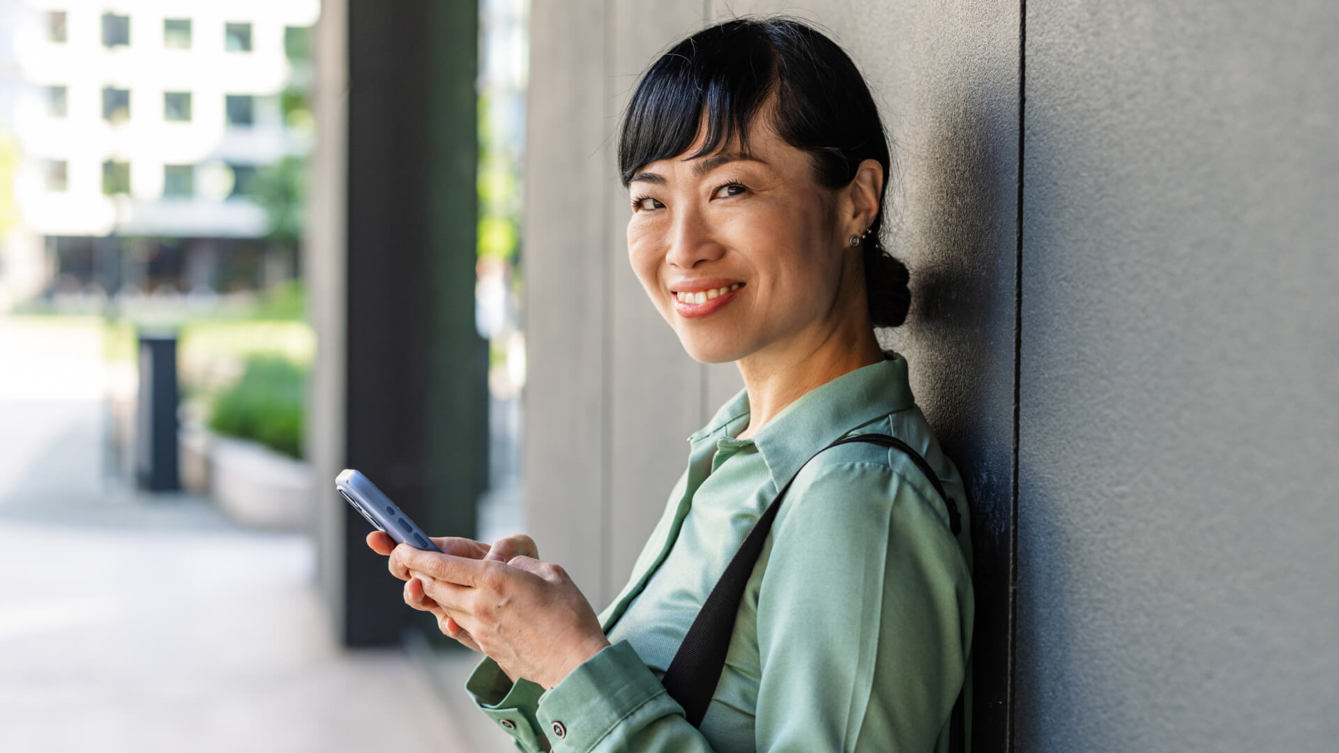 Person standing outside against a modern building wall, wearing a green shirt and shoulder bag, holding a smartphone in both hands.