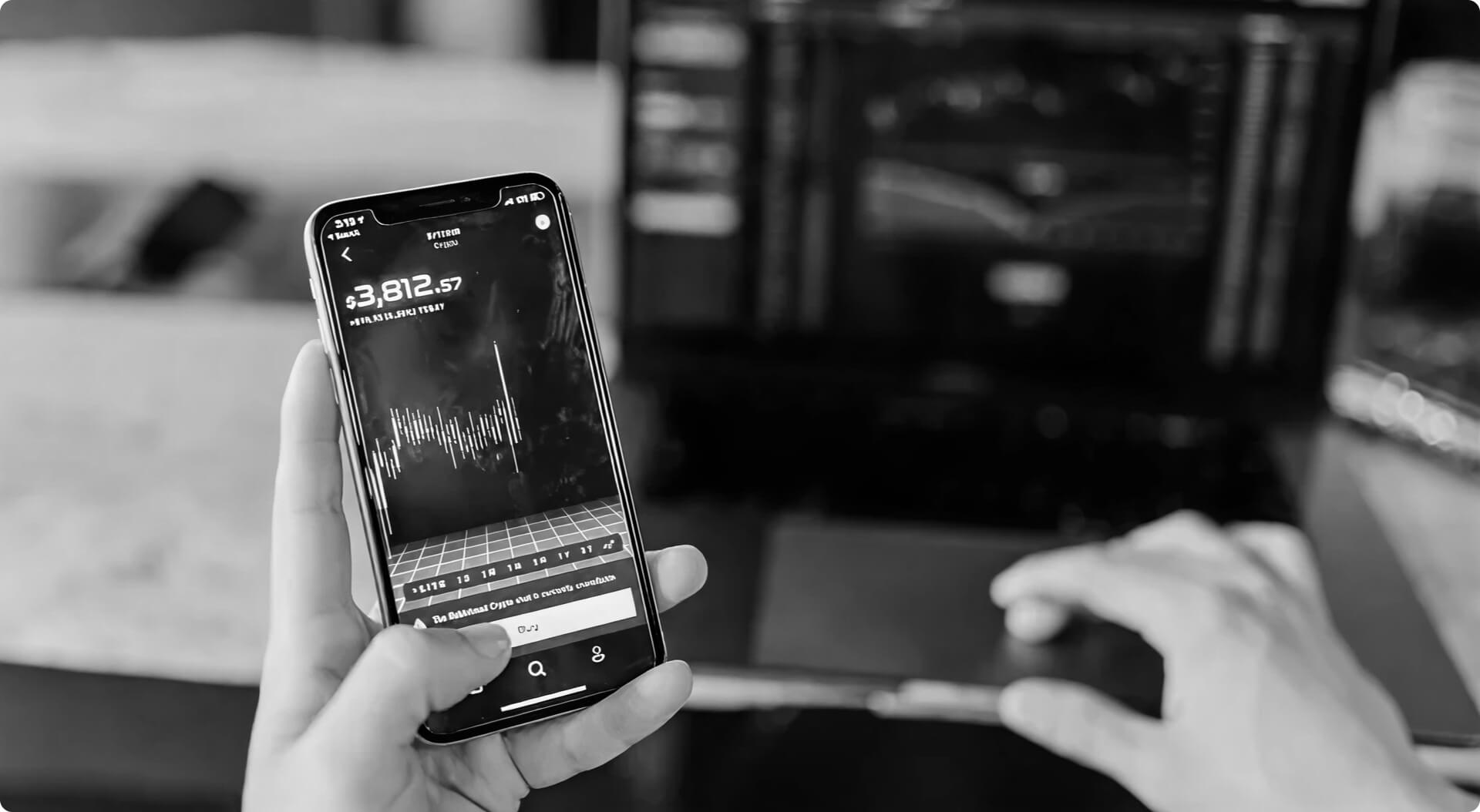 Grayscale close-up of a hand holding a smartphone with financial charts on the screen in front of a blurred laptop showing data graphs.