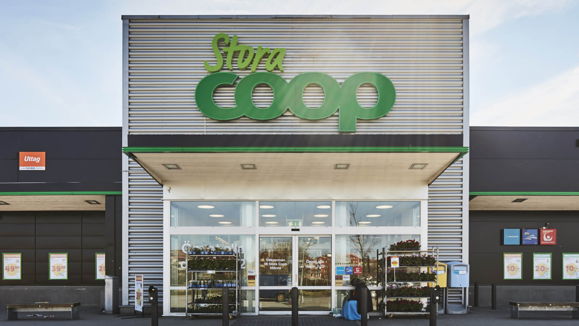 Front view of a Coop supermarket entrance with large green logo, sliding glass doors, and outdoor displays of plants and products.