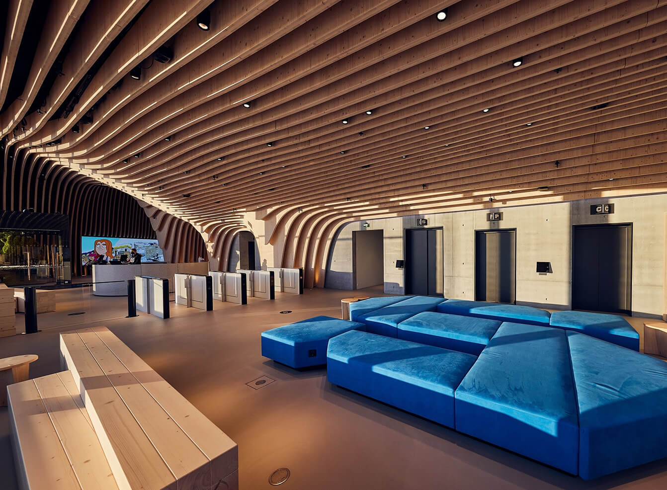 Wide shot of a modern lobby with a sculpted wooden ceiling, blue modular sofas, turnstiles, and a wall of elevators in warm lighting.