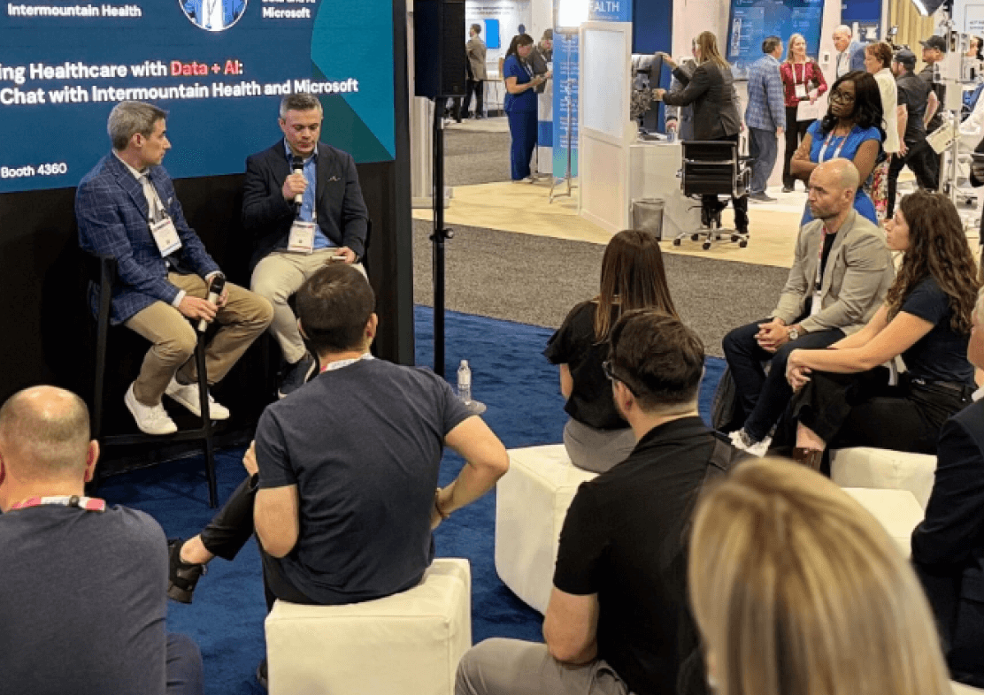 Audience seated on stools listening to a small panel discussion at a busy conference booth.