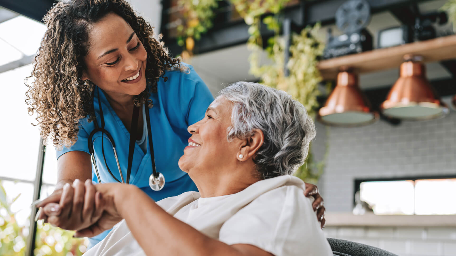 Nurse in blue scrubs holding hands and talking with an older patient in a bright, modern care setting.