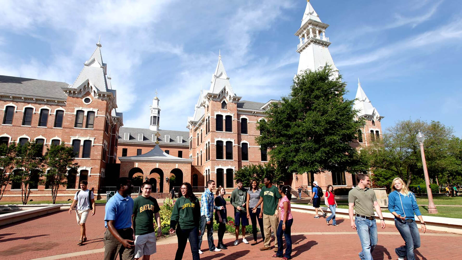 Students walking in front of red-brick Baylor University campus buildings under a bright blue sky.