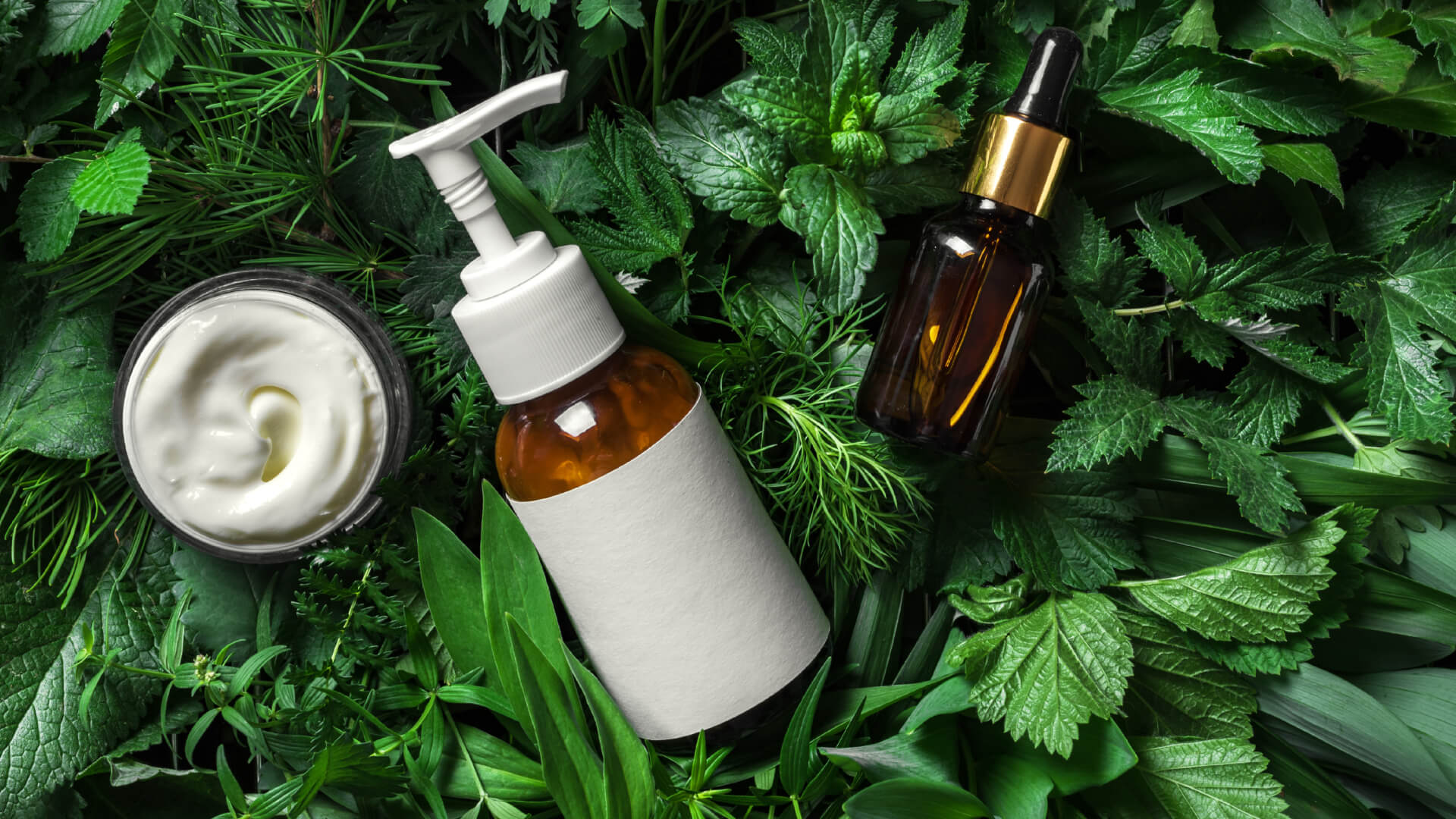 Top-down view of natural skincare products, including a cream jar and two amber bottles, arranged on a bed of fresh green leaves and herbs.