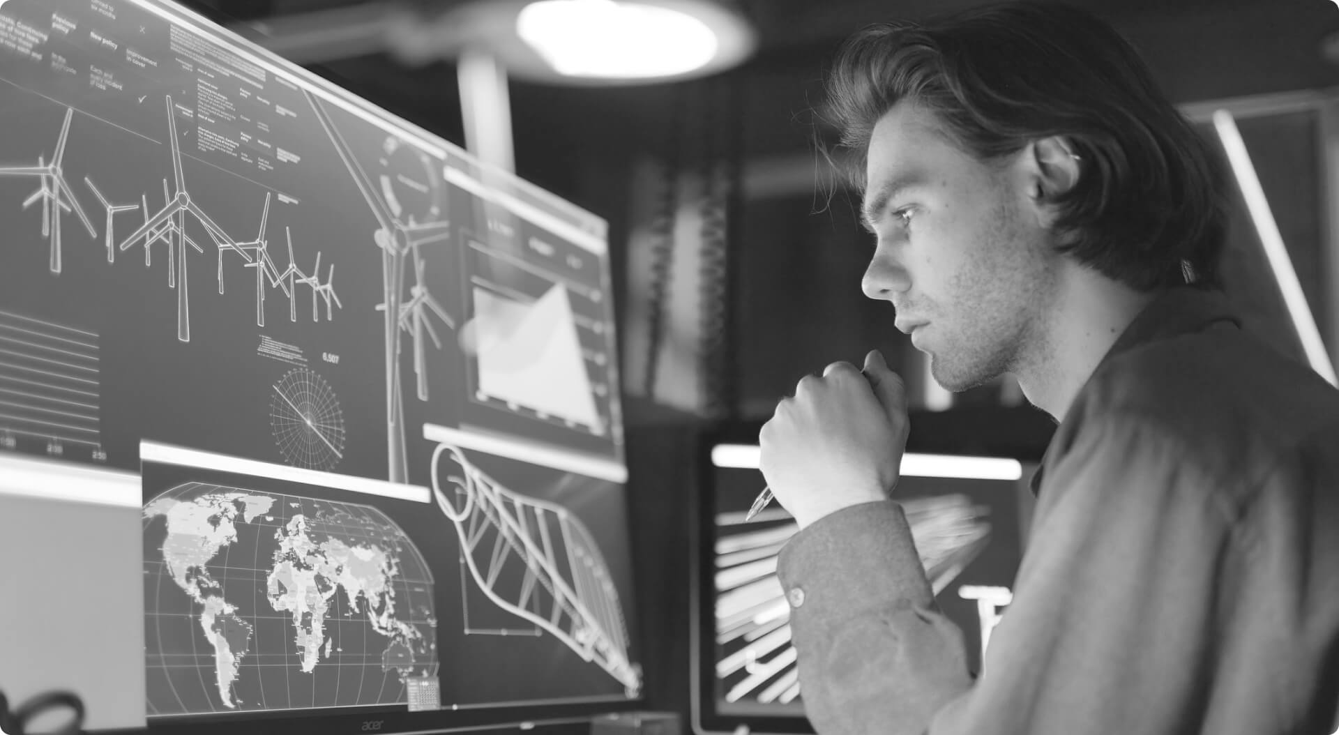 Grayscale photo of a person studying multiple large data visualization screens in a dark workspace.
