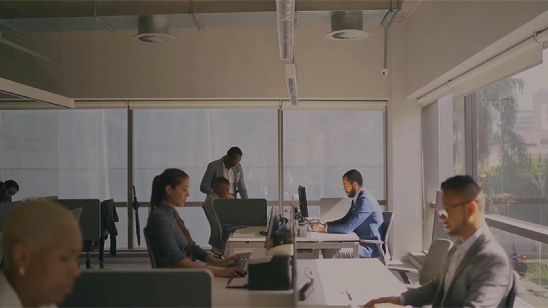 Office scene with employees working at desks in a bright, modern workspace with large windows.