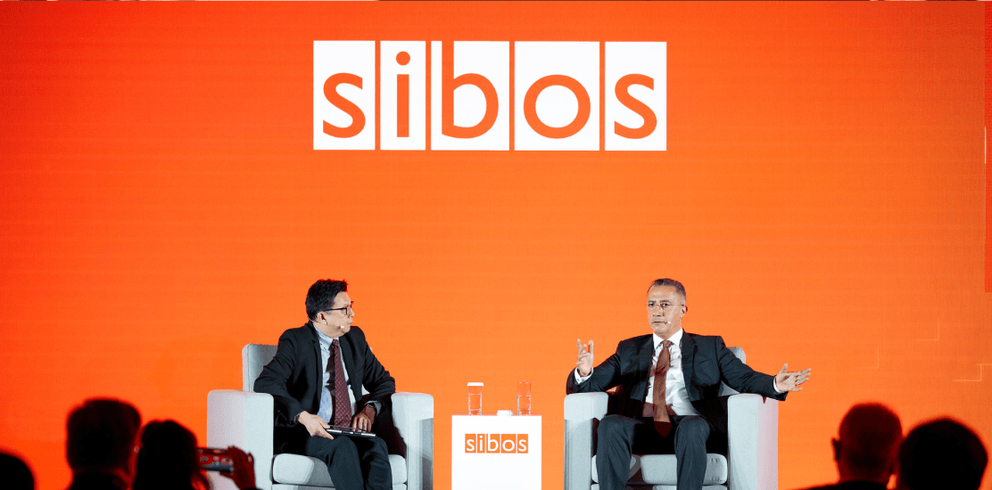 Two men sit on a stage under a Sibos sign.