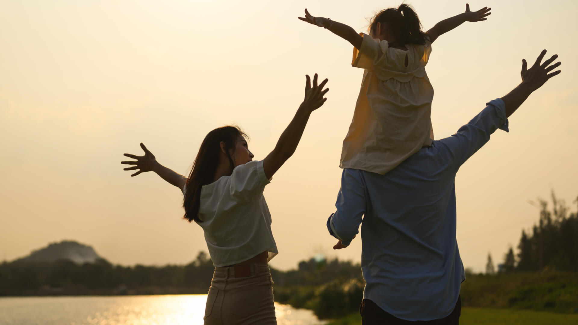 Family with child on shoulders, arms outstretched at sunset.