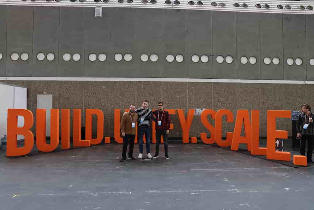 Three attendees pose in front of large orange physical letters that spell out "Build. Unify. Scale." behind them at Spark + AI Summit 2019.